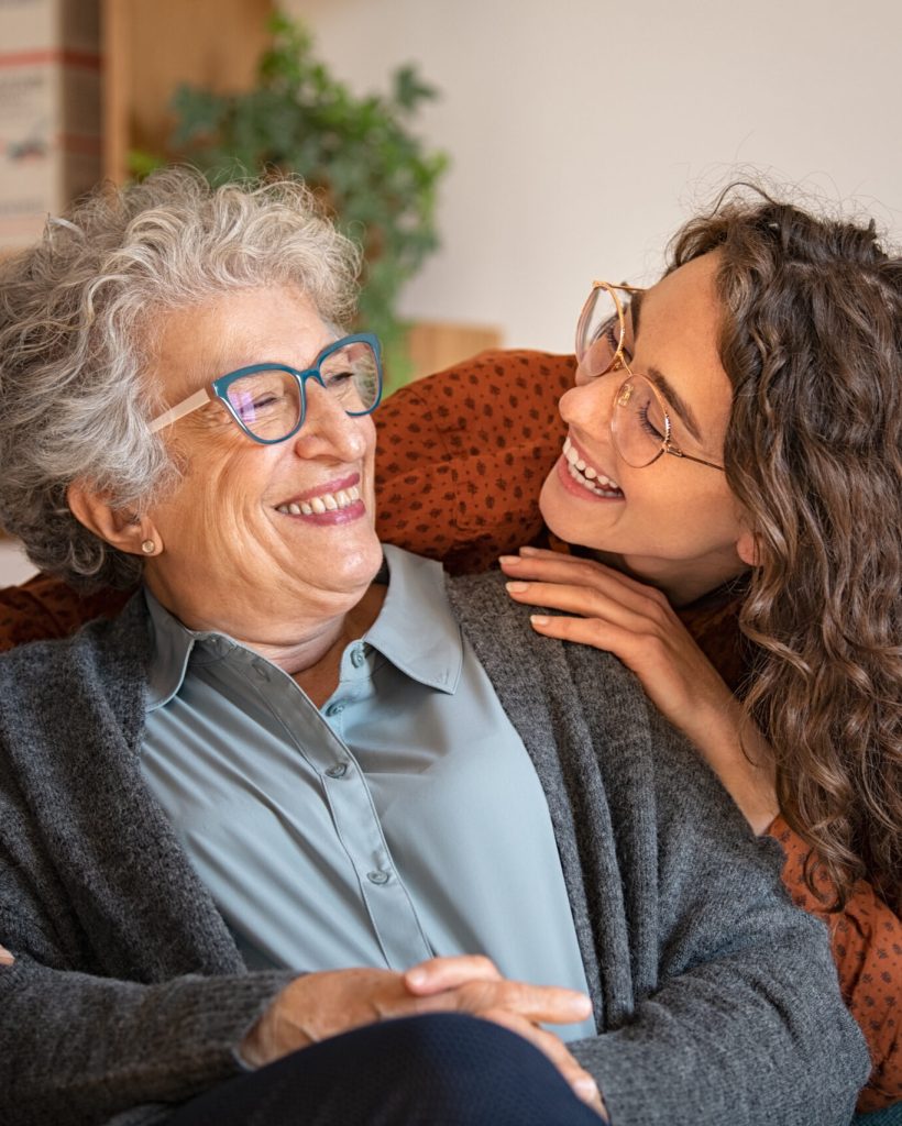 Old grandmother and adult granddaughter hugging at home and looking at each other. Happy senior mother and young daughter embracing with love on sofa. Happy young woman hugging from behind grandma with love.