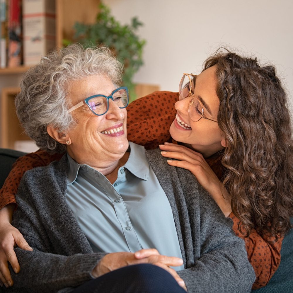 Old grandmother and adult granddaughter hugging at home and looking at each other. Happy senior mother and young daughter embracing with love on sofa. Happy young woman hugging from behind grandma with love.